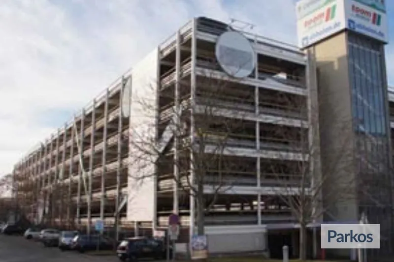 Mehrstöckiges Parkhaus mit offenen Parkebenen an einer Straße, davor geparkte Autos und kahle Bäume.