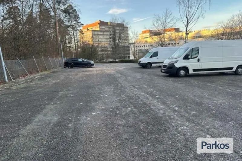 Schotterparkplatz mit zwei weißen Transportern und einem schwarzen Auto, Gitterzaun links, Bürogebäude und Bäume im Hintergrund.