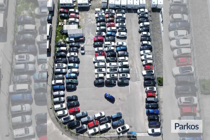 Luftaufnahme eines umzäunten Parkplatzes mit dicht geparkten Reihenfahrzeugen und blauem Wagen in der Mitte.