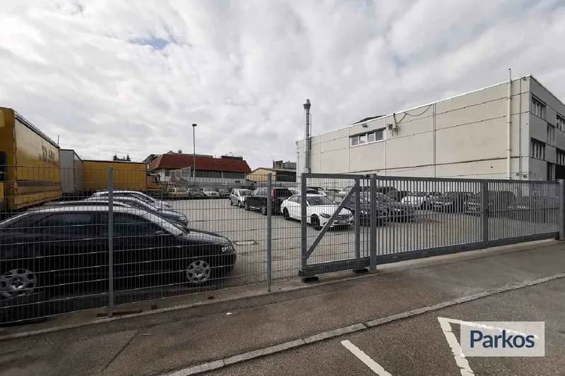Eingezäunter Parkplatz mit geöffnetem Schiebetor, geparkten Autos und einem Industriegebäude im Hintergrund.
