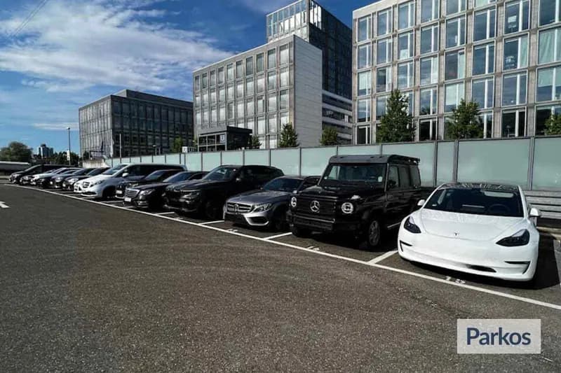Reihe geparkter Autos, darunter weißer Tesla und schwarzer Mercedes G-Klasse, auf eingezäunter Freifläche vor Bürogebäuden.