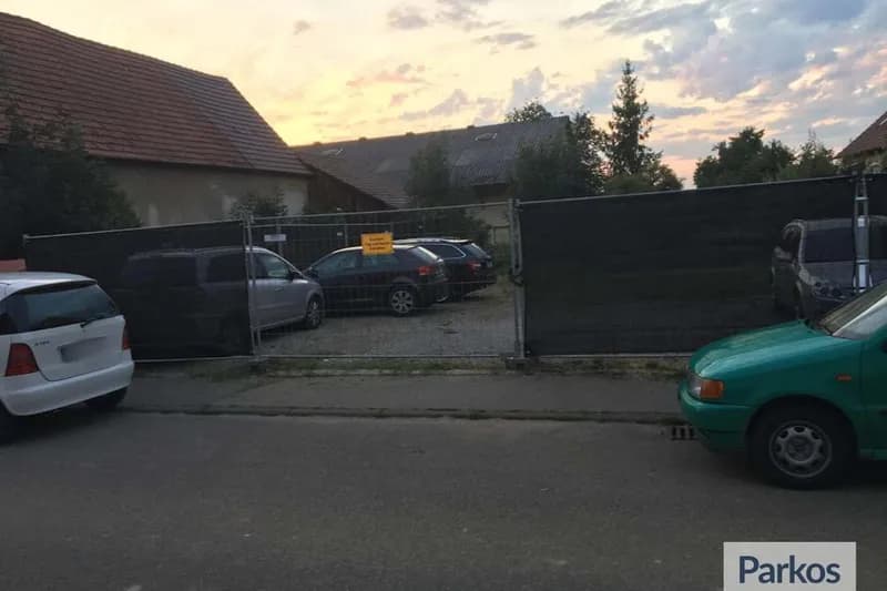 Umzäunter Schotterparkplatz mit mehreren geparkten Autos hinter Bauzaun und Sichtschutz, Häuser im Hintergrund.