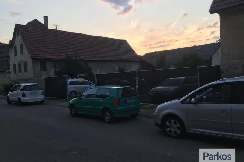Geparkte Autos an einer Straße vor einem eingezäunten Freigelände und Wohnhäusern bei Sonnenuntergang.