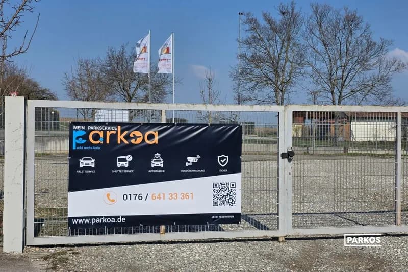 Metalltor mit Parkoa-Werbeplanen am umzäunten Schotterparkplatz, Fahnen und Bäume im Hintergrund.