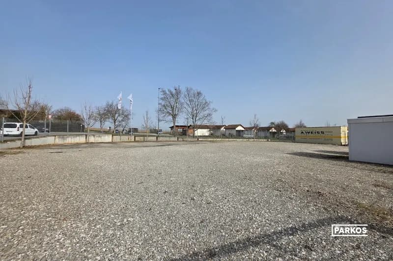 Große kiesige Freifläche mit Umzäunung, Fahnen, Containern und vereinzelten Bäumen unter klarem Himmel.