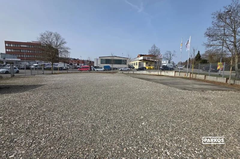 Umzäunter Schotterparkplatz mit Fahnen, Gebäuden und geparkten Fahrzeugen im Hintergrund.