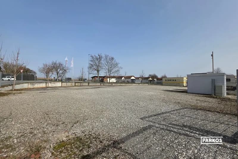 Eingezäunter Schotterparkplatz mit einzelnen Bäumen, Containern und Fahnen unter blauem Himmel.