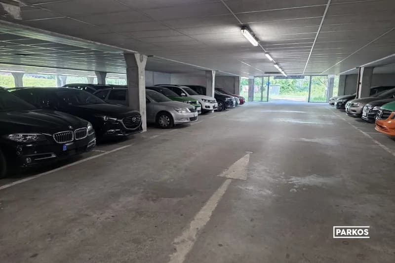 Überdachtes Parkdeck mit geparkten Autos links und rechts, Betonpfeil auf dem Boden, Stützsäulen und Gitterzaun im Hintergrund.