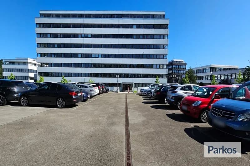Freifläche mit Fahrspur und parkenden Autos links und rechts vor mehrstöckigem Bürogebäude bei blauem Himmel.