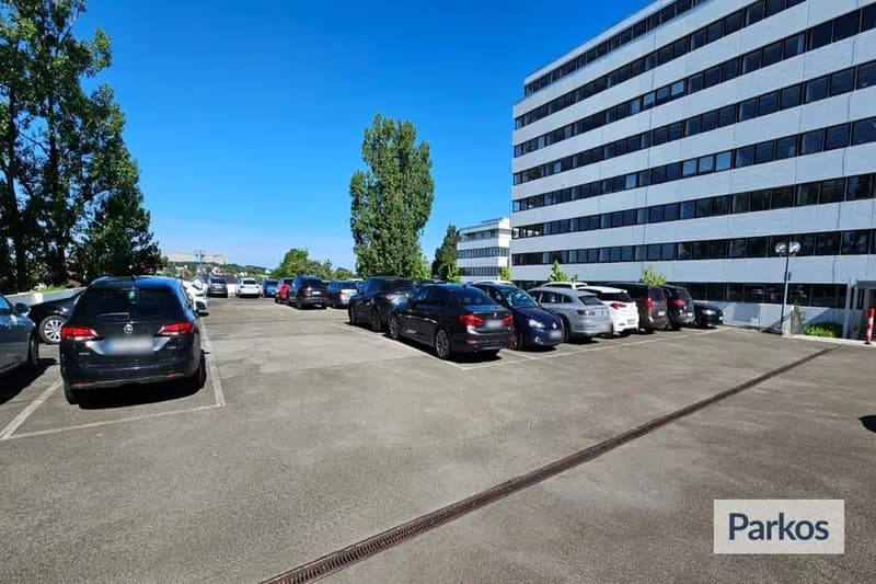 Freifläche mit geparkten Autos auf Asphalt neben einem mehrstöckigen Bürogebäude und Bäumen unter blauem Himmel.