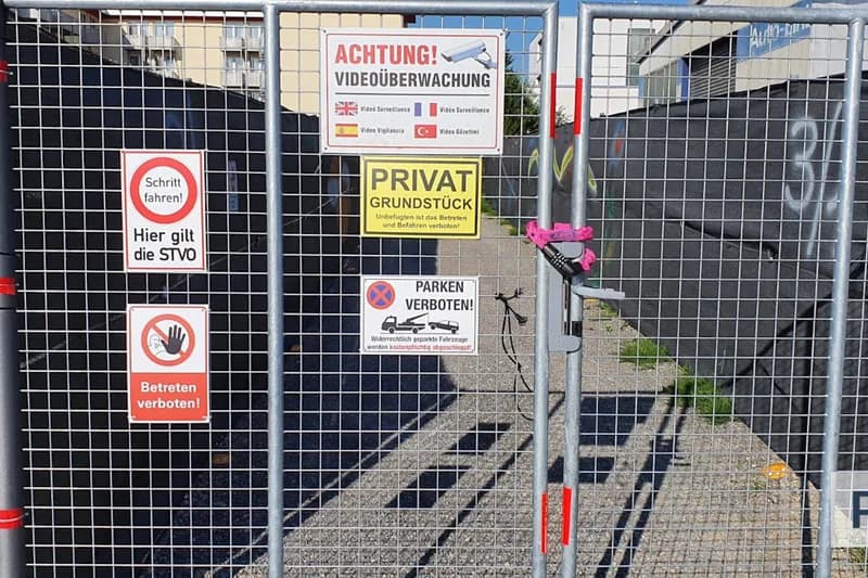 Metallgittertor mit Vorhängeschloss und mehreren Verbotsschildern wie Videoüberwachung, Privatgrundstück und Parken verboten; Kiesweg sichtbar.