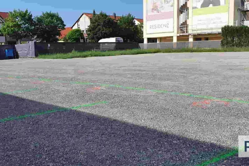 Leerer geschotterter Parkplatz mit grünen Stelllinien, roten Markierungen, Zaun und Werbetafel im Hintergrund.