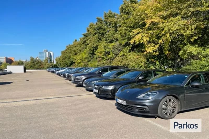 Reihe dunkel lackierter Autos auf offenem Parkplatz vor Baumreihe und blauem Himmel, Parkos-Logo unten rechts.