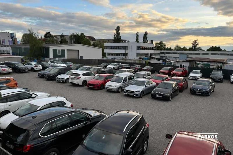 Große Schotterparkfläche mit mehreren Reihen geparkter Autos, Betriebsgebäude und wolkiger Abendhimmel.