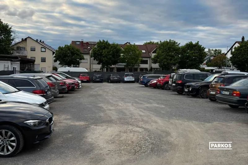 Geschotterter Parkplatz mit zwei Reihen geparkter Autos und Wohnhäusern im Hintergrund.