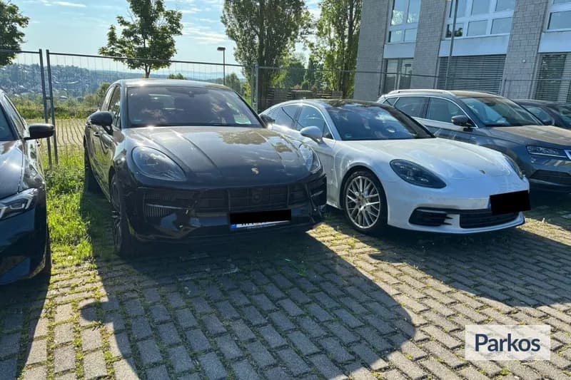 Zwei geparkte Porsche nebeneinander auf gepflasterter Freifläche vor Zaun und Gebäude.