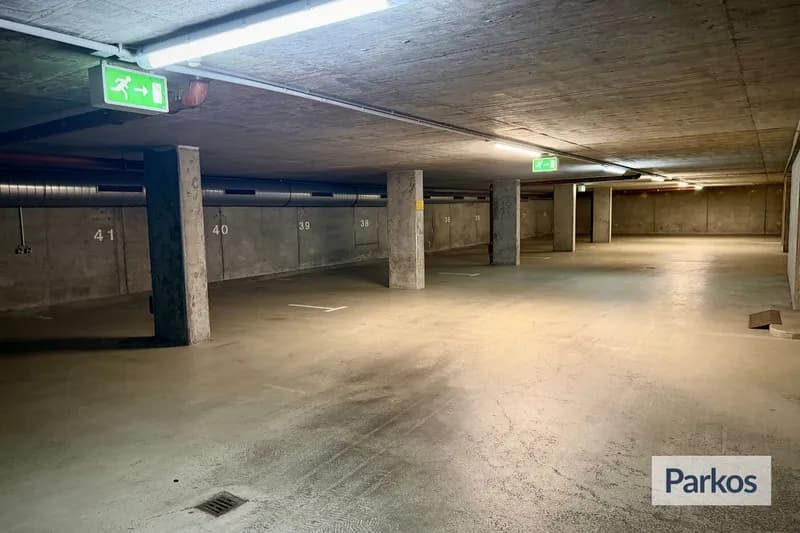 Überdachtes Tiefgaragenparkdeck mit markierten Stellplätzen, Betonstützen und grünen Notausgangsschildern.