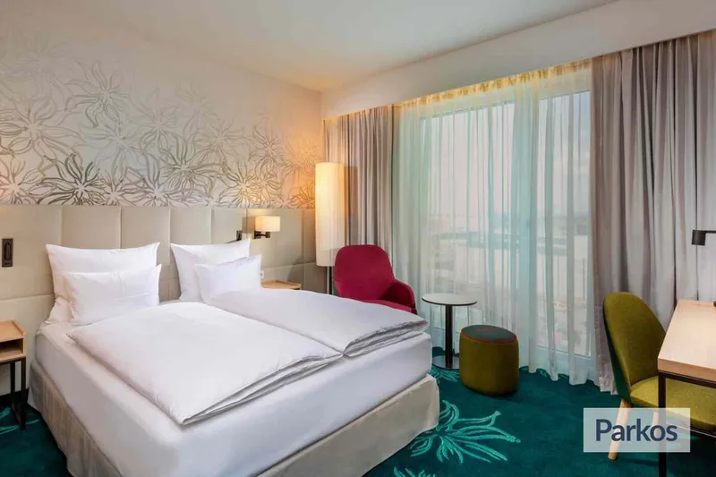Hotelzimmer mit großem Doppelbett, gepolstertem Kopfteil, roten und grünen Sesseln sowie bodentiefem Fenster mit Vorhängen.