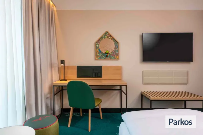 Hotelzimmer mit Schreibtisch, grünem Stuhl, Wandspiegel, Fernseher, Bettkante und Gardine.