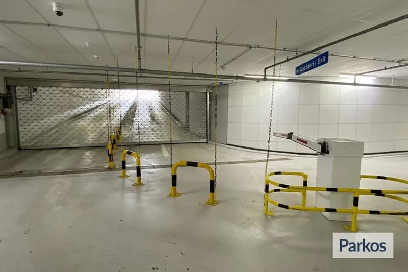 Überdachtes Parkhaus mit geschlossenem Metallgittertor, geöffnetem Schrankenarm und gelb-schwarzen Pollern und Ketten.