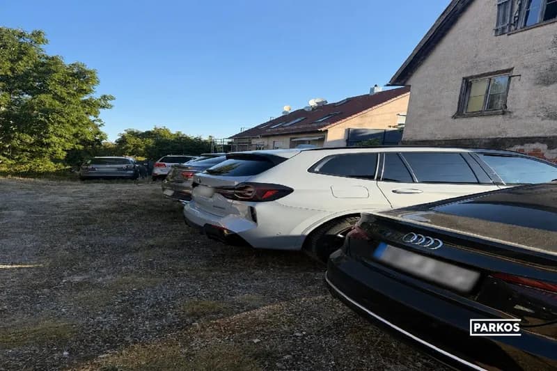 Mehrere geparkte Autos auf einem kiesigen Freiflächenparkplatz neben Wohnhäusern.