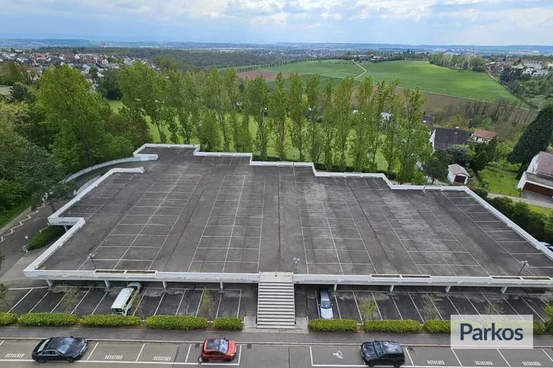 Flaches Parkdeck mit markierten Stellflächen, mittiger Treppe zur Straße und grüner Landschaft im Hintergrund.